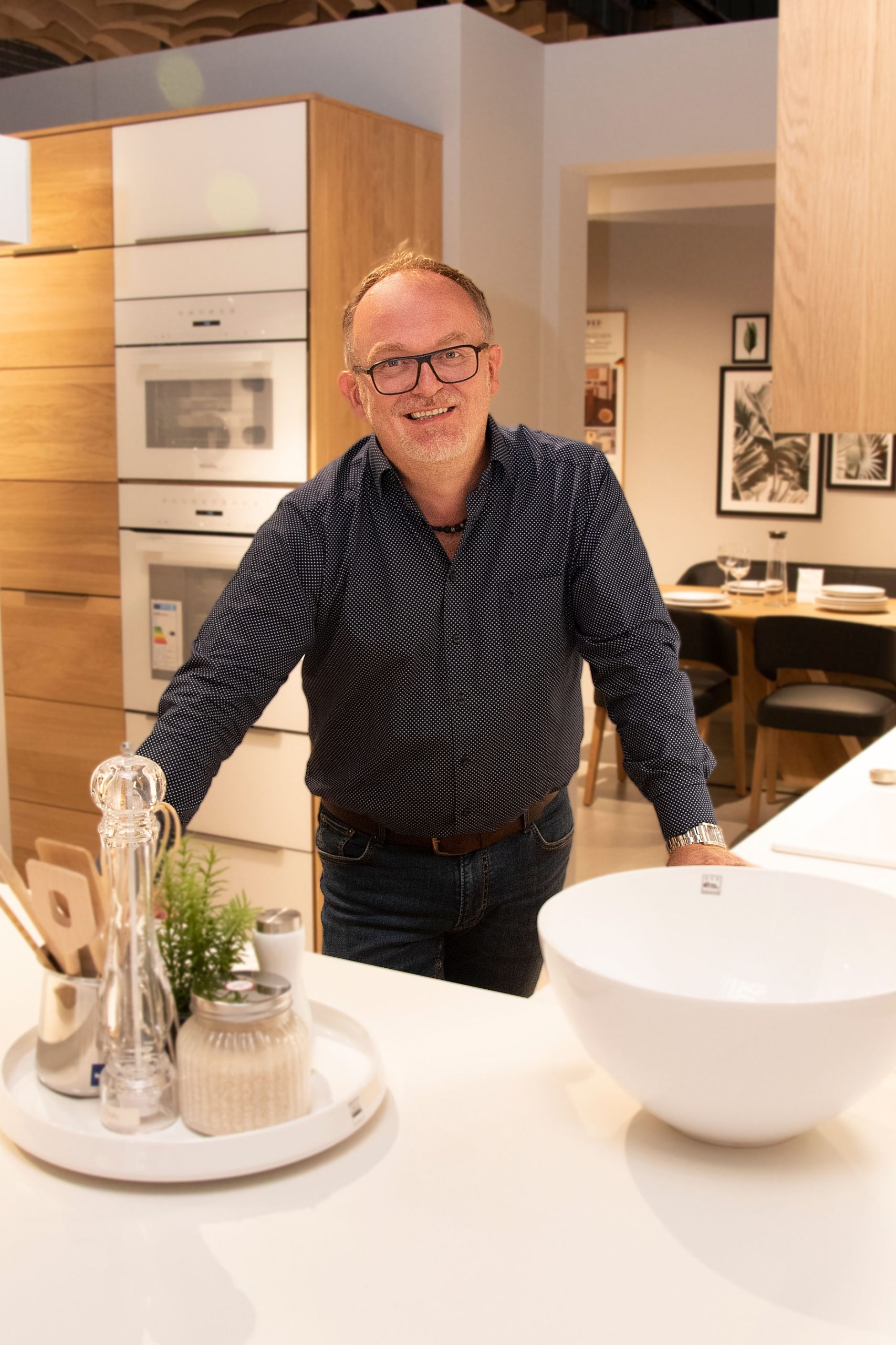 Ein Mann mit Brille lächelt, als er in einer modernen Küche neben einer weißen Arbeitsplatte mit Dekorationsartikeln steht.