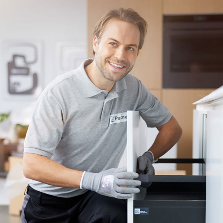 Ein lächelnder Techniker in einem grauen Polohemd und Handschuhen repariert ein modernes Küchengerät in einem Haus.