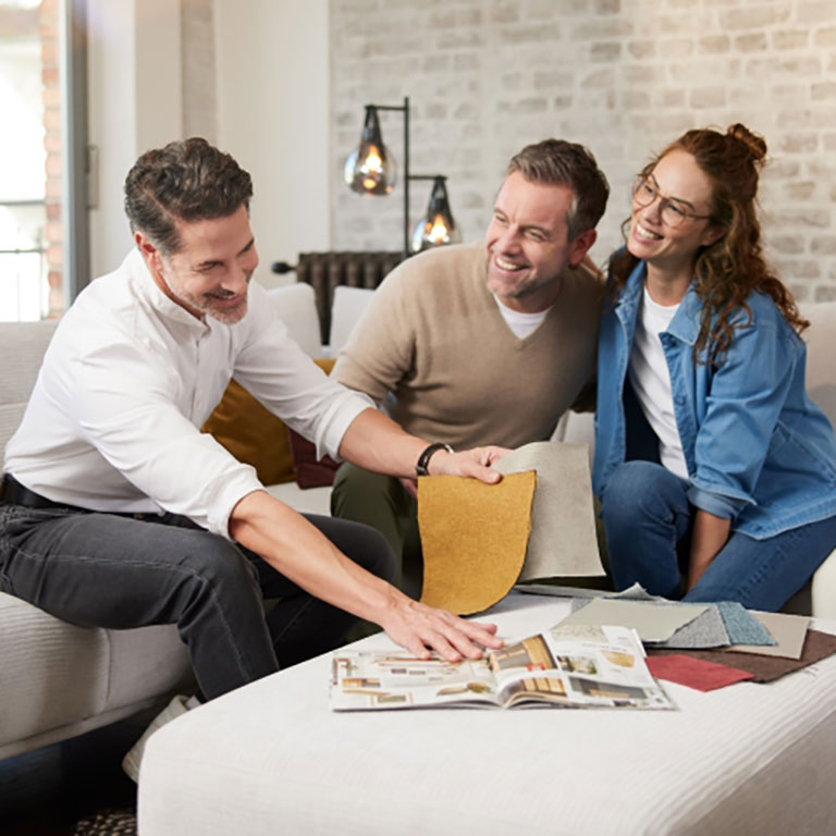 Drei Personen sitzen auf einer Couch, lächeln und betrachten Stoffmuster und eine Zeitschrift mit Einrichtungsideen.
