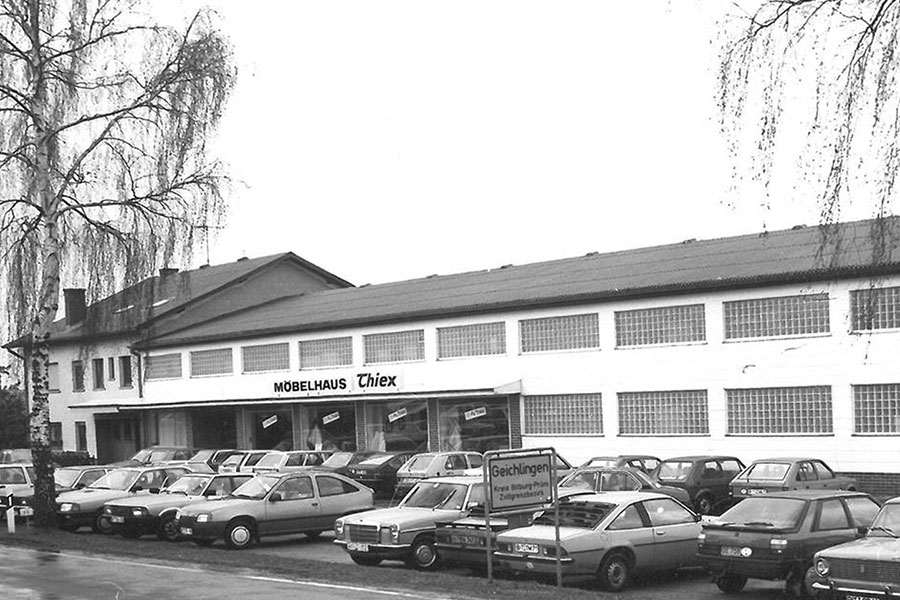 Schwarz-Weiß-Foto des Möbelhauses Thiex mit vielen geparkten Autos und Bäumen vor dem Gebäude.