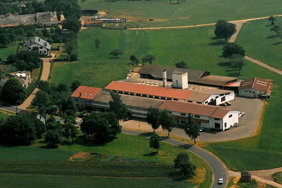 Luftaufnahme eines großen Bauernhofkomplexes mit rotgedeckten Gebäuden, umgeben von grünen Feldern und Bäumen.