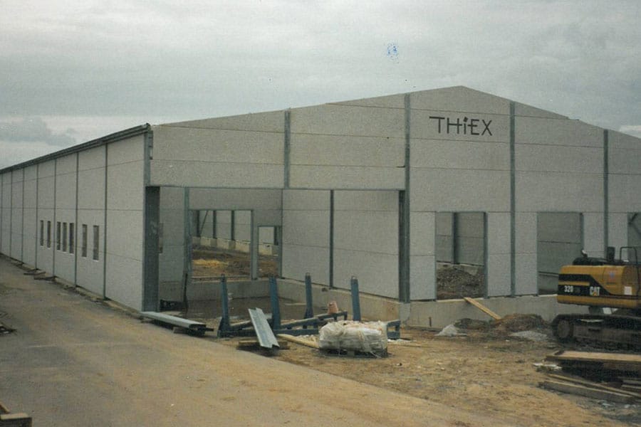 Ein großes Industriegebäude im Bau mit THIEX auf der Fassade und einem gelben Bagger in der Nähe.