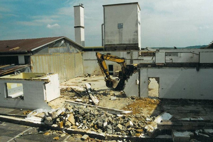 Ein gelber Bagger reißt ein Gebäude ab; Schutt und Trümmer liegen auf der Baustelle verstreut.