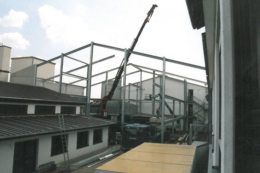 Ein Kran hebt Stahlträger auf einer Baustelle an, die von teilweise fertiggestellten Gebäuden umgeben ist.