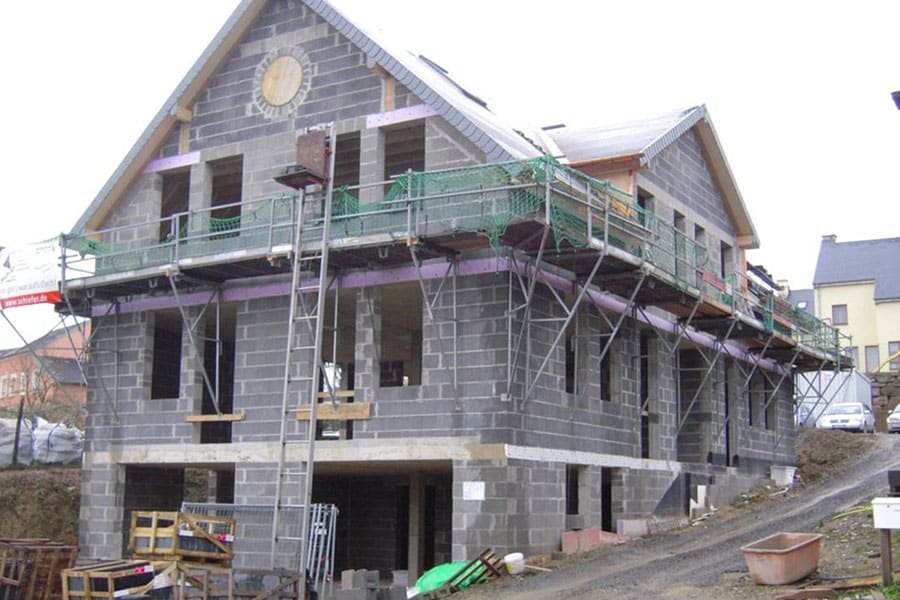 Ein zweistöckiges Haus im Bau mit Gerüsten und Leitern an der Außenseite, gebaut aus grauen Betonblöcken.