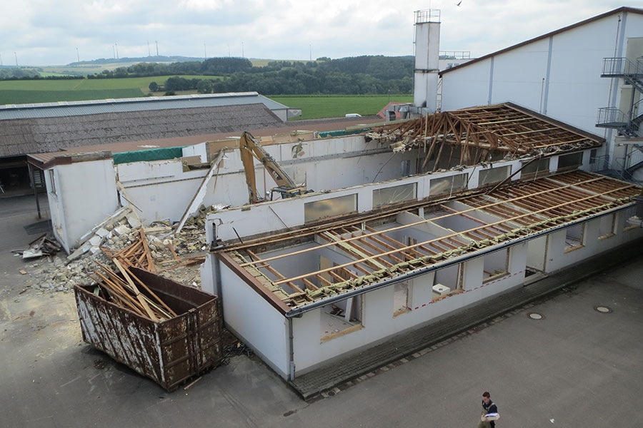 Ein teilweise abgerissenes Gebäude mit Trümmern und freiliegenden Holzbalken neben einem großen Müllcontainer in einem Industriegebiet.