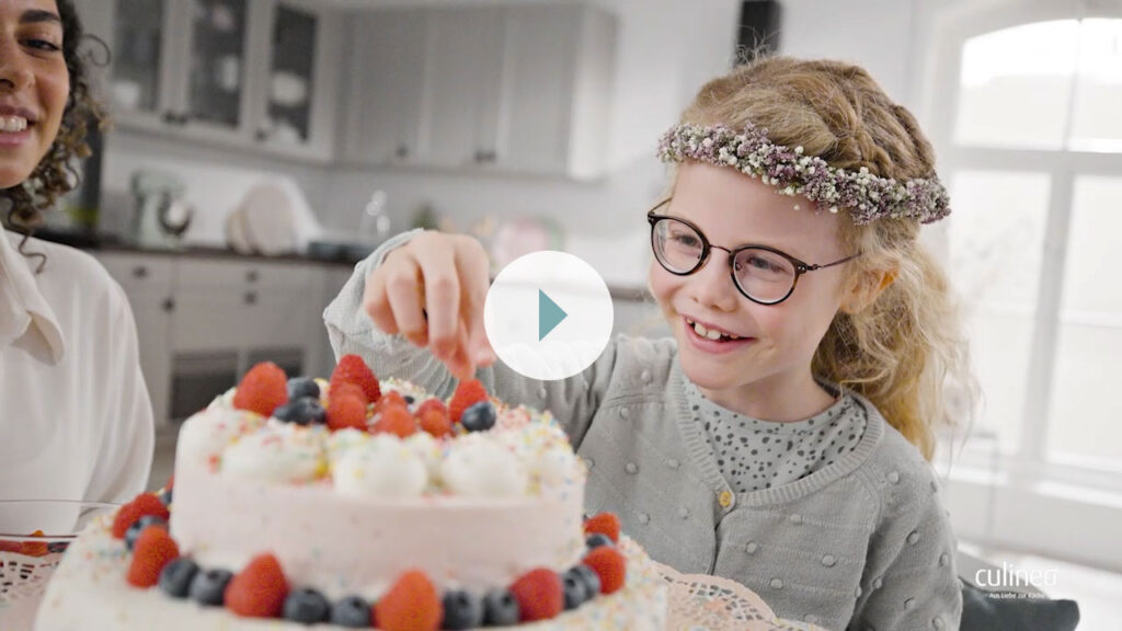 Ein lächelndes Mädchen mit Brille dekoriert einen Kuchen mit Beeren in einer hellen Küche und trägt ein geblümtes Stirnband.