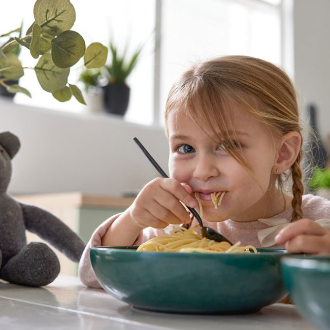 Ein junges Mädchen mit geflochtenen Haaren isst lächelnd Spaghetti aus einer grünen Schüssel und sitzt neben einem grauen Plüschbären.