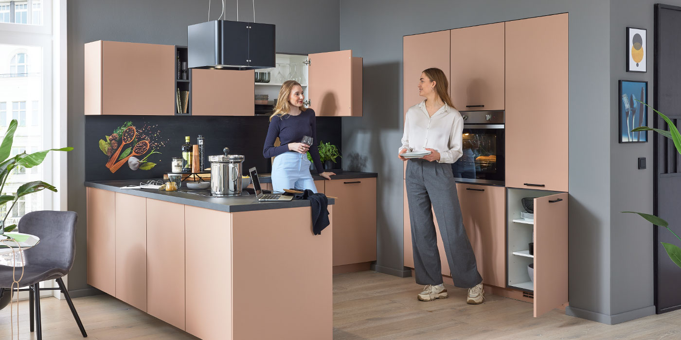 Zwei Frauen plaudern und lächeln in einer modernen Küche mit hellbraunen Schränken und schwarzen Arbeitsplatten.