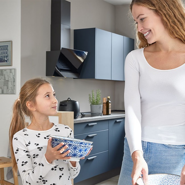 Eine Frau und ein Mädchen lächeln sich in einer modernen Küche an und halten gemusterte Schüsseln.