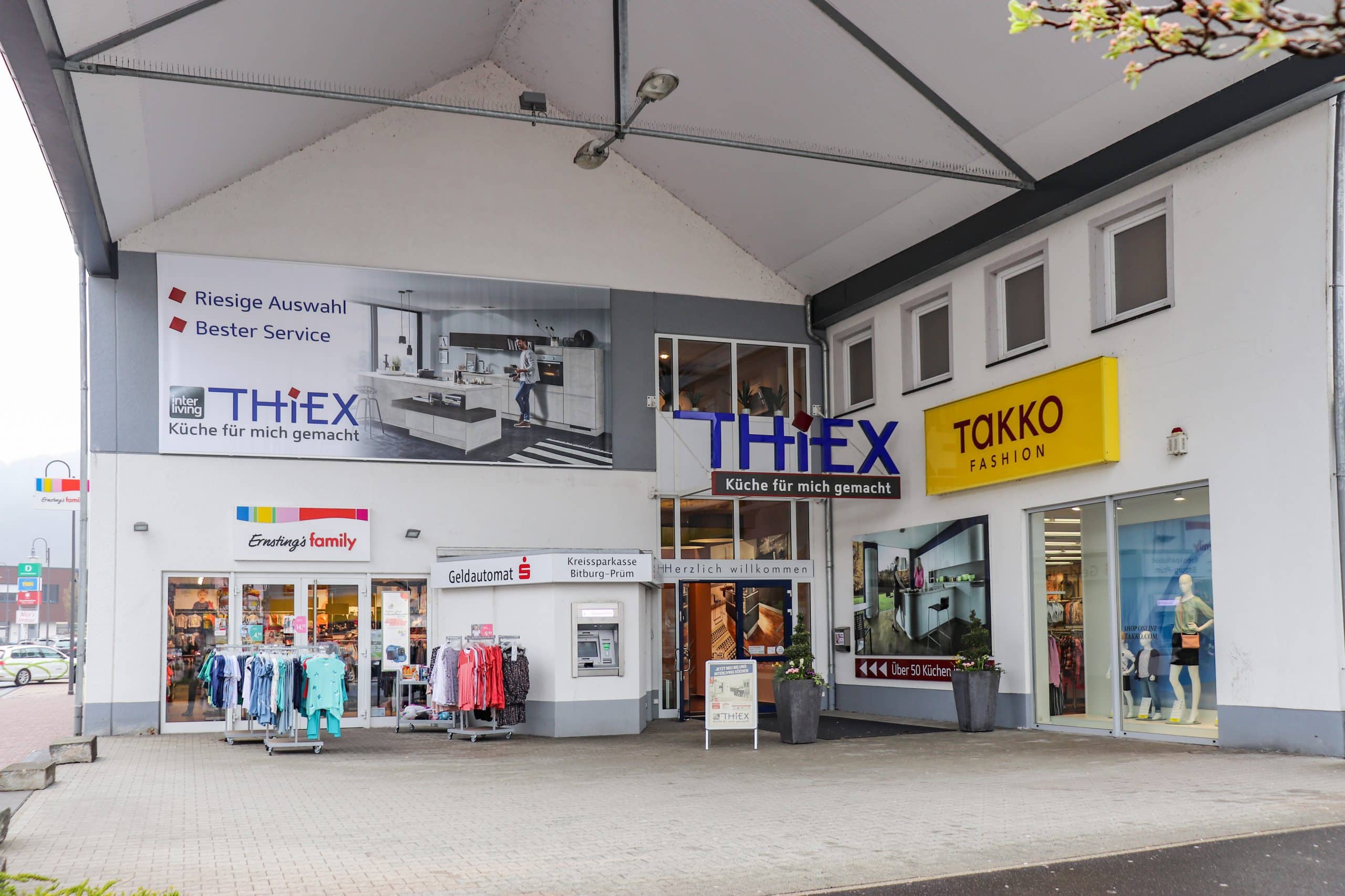 Schaufenster von Thiex Küchen und Takko Fashion mit Kleiderständern und einem Geldautomaten im Freien unter einem überdachten Steg.
