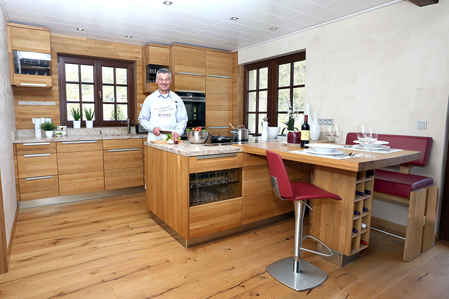 Ein Mann bereitet in einer modernen Holzküche an einer Insel mit roten Stühlen und einem Tisch für zwei Personen das Essen zu.