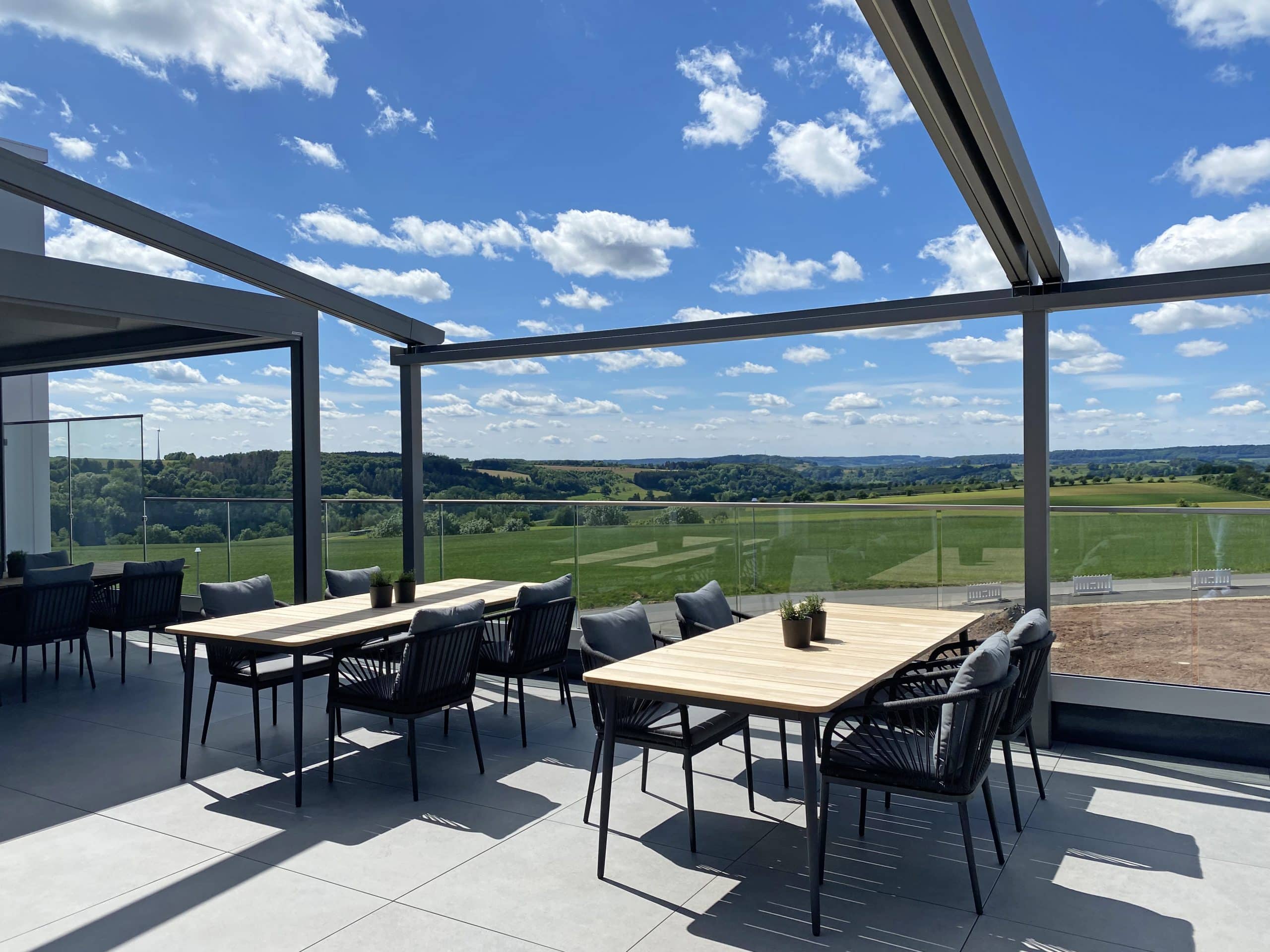Außenterrasse mit Tischen und Stühlen mit Blick auf grüne Felder und Hügel unter einem blauen Himmel mit vereinzelten Wolken.