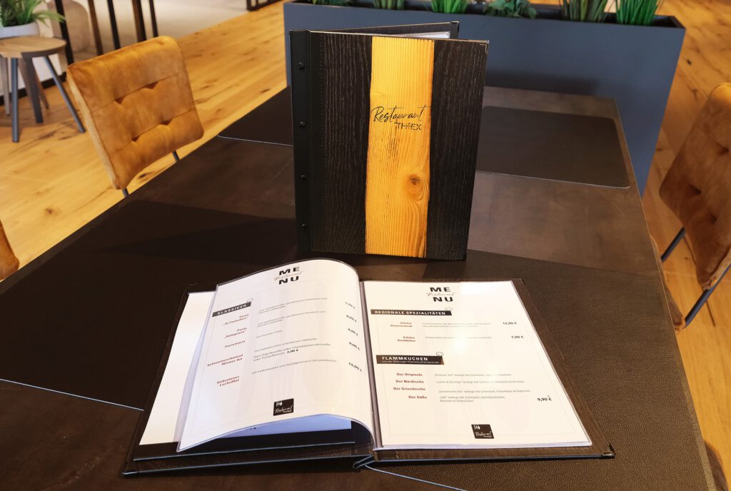 Zwei offene Speisekarten und eine geschlossene Speisekarte liegen auf einem dunklen Tisch in einem modernen Restaurant mit braunen Stühlen.
