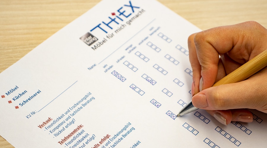 Eine Hand, die einen Stift in der Hand hält, füllt ein Bewertungsformular für Möbel mit Kästchen und deutschem Text aus.