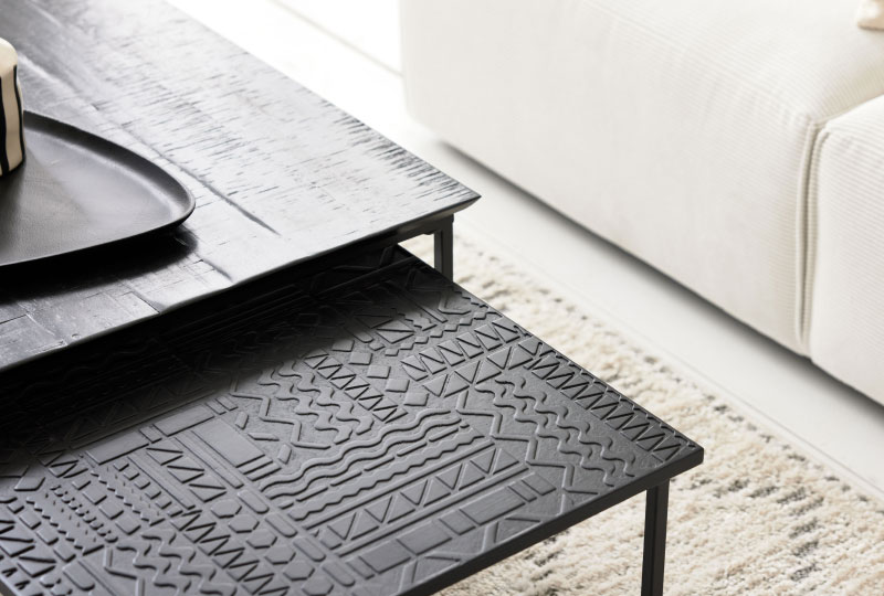 Nahaufnahme von zwei schwarzen Nisttischen mit geometrisch strukturierten Oberflächen neben einem cremefarbenen Sofa und einem gemusterten Teppich.