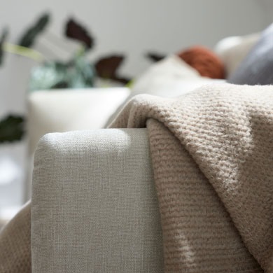 Nahaufnahme einer beigen Strickdecke, die über den Arm eines hellen Sofas drapiert ist, mit unscharfen Pflanzen im Hintergrund.