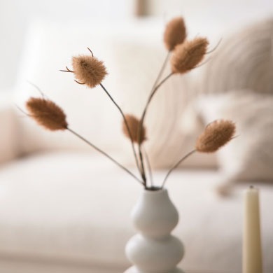 Getrocknete braune Gräser in einer weißen Vase auf einem Tisch mit einer verschwommenen beigen Couch im Hintergrund.