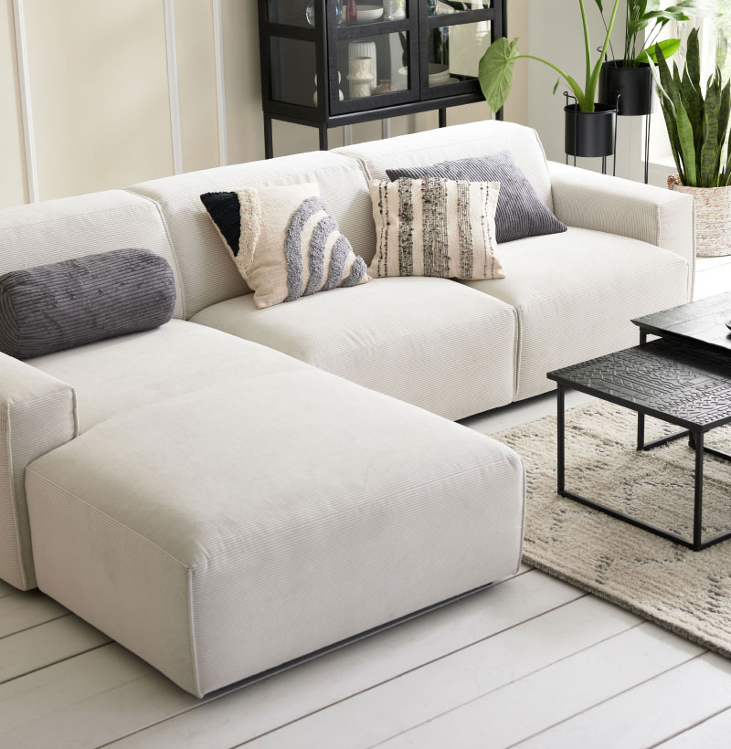 Modernes Wohnzimmer mit einem weißen Sektionssofa, dekorativen Kissen, einem schwarzen Tisch und mehreren Grünpflanzen.
