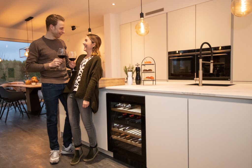 Ein lächelndes Paar steht in einer modernen Küche, hält Gläser mit Rotwein in der Hand und sieht sich an.