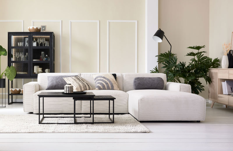 Modernes Wohnzimmer mit weißem Sektionssofa, schwarzen Tischen, Pflanzen und minimalistischem Dekor in neutralen Tönen.