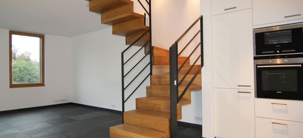 Moderne Küche mit Einbaugeräten und Holztreppe mit schwarzem Geländer, in der Nähe eines großen Fensters.