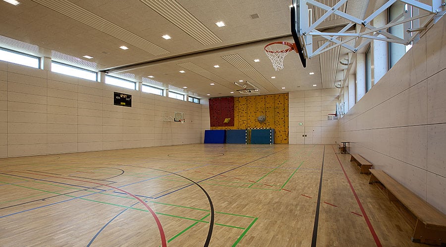Leere Turnhalle mit Basketballkörben, Holzboden, Bänken und einer Kletterwand am anderen Ende.