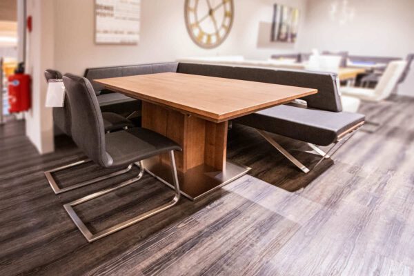 Eckbankgruppe Vista 100: Holztisch, schwarze Stühle und Eckbank auf dunklem Holzboden im Ausstellungsraum.