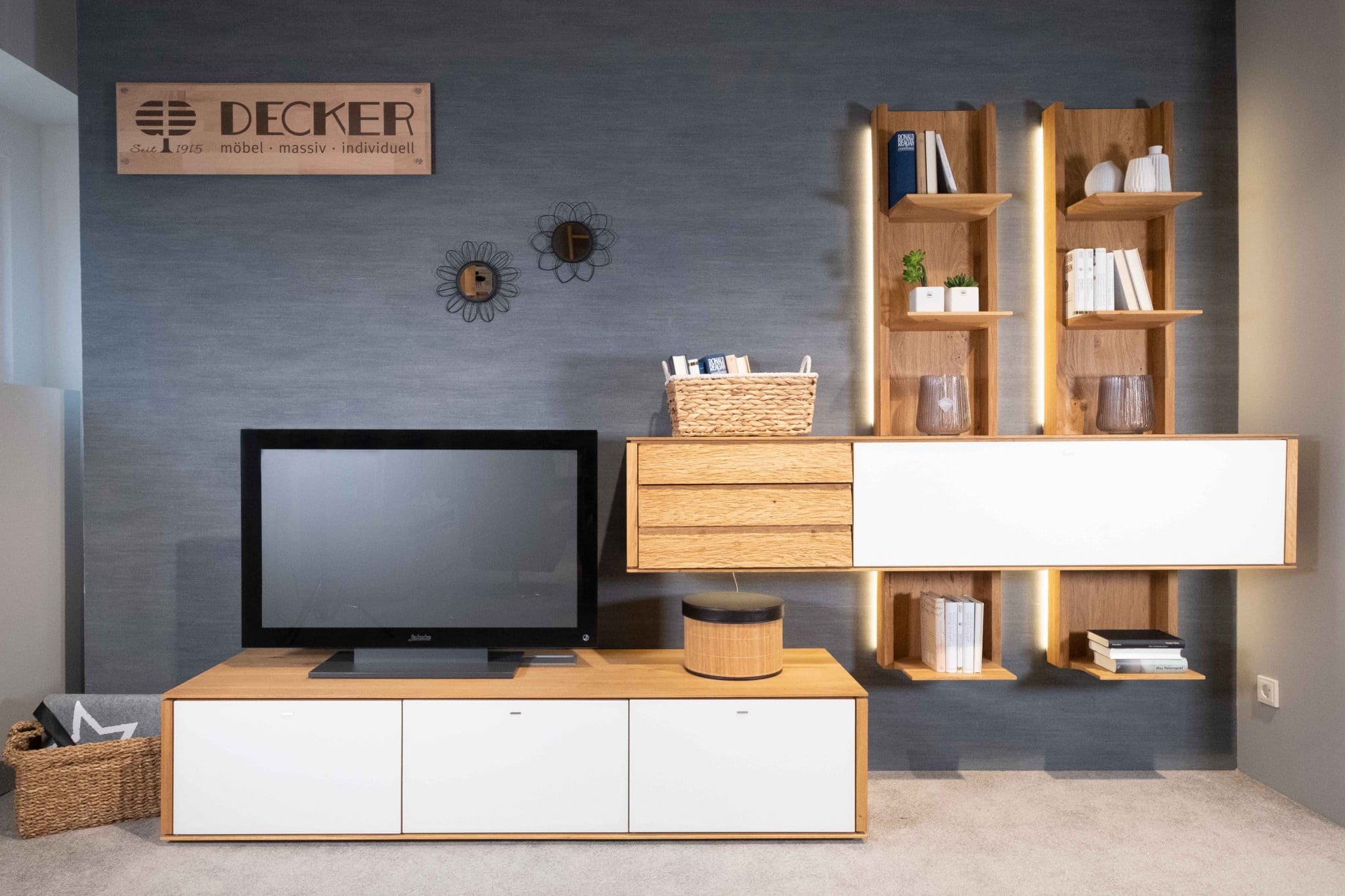 Wohnprogramm Alivio: Modernes Wohnzimmer mit TV, Holzschränken, Regalen, Büchern und Dekoration an einer grau strukturierten Wand.
