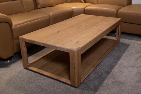 Der Couchtisch Sapio 9500, ein rechteckiger Holztisch mit unterer Ablage, steht auf grauem Teppich neben einem hellbraunen Ledersofa.