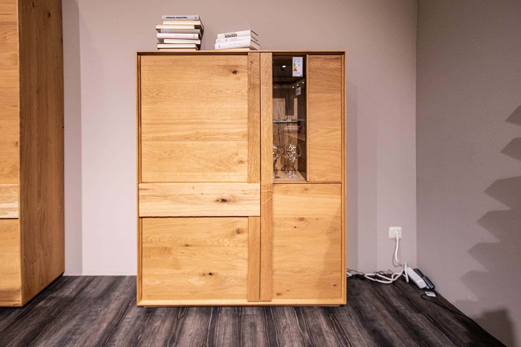 Highboard Woodline mit gestapelten Büchern darauf, Gläsern darin und Stromkabeln daneben auf dem Boden.