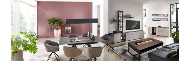 Die Wohnkoje Alva verfügt über einen modernen Ess- und Wohnbereich, neutrale Möbel, große Fenster, Pflanzen und eine malvenfarbene Akzentwand.