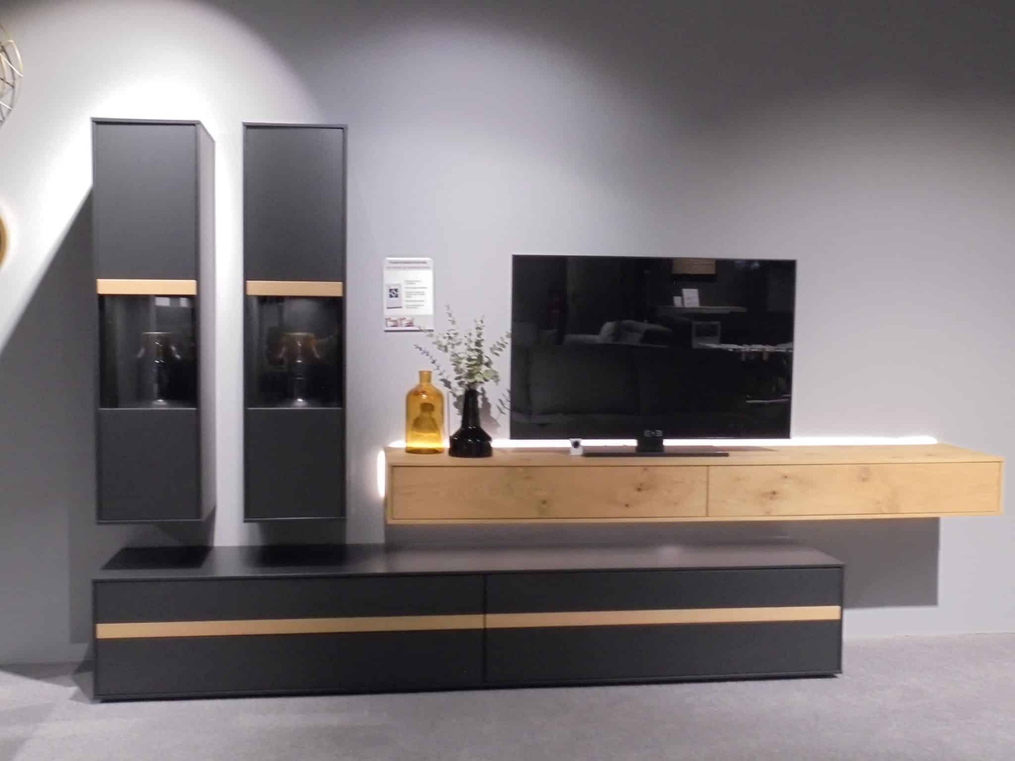 Wohnprogramm Calea TV-Element mit schwarzen und hölzernen Schränken, montiertem TV und Vasen an einer grauen Wand.