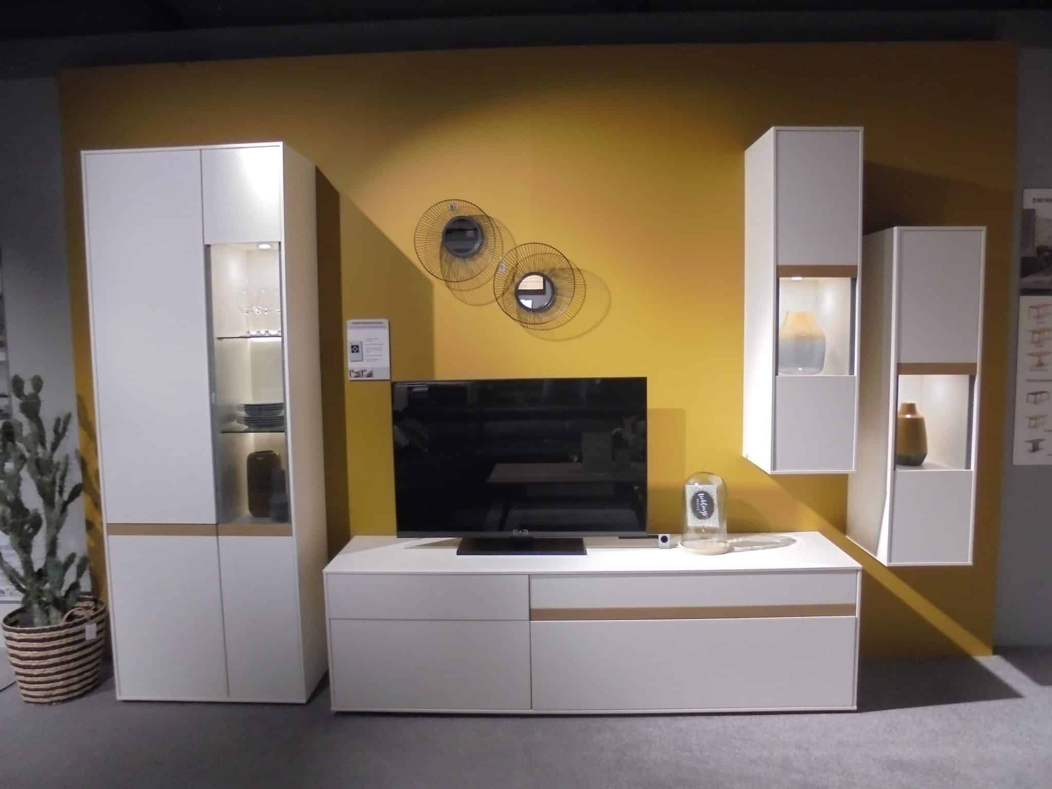 Wohnprogramm Calea: Moderner weißer TV-Schrank, gelber Wandhintergrund, Regale und zentraler schwarzer TV-Bildschirm.
