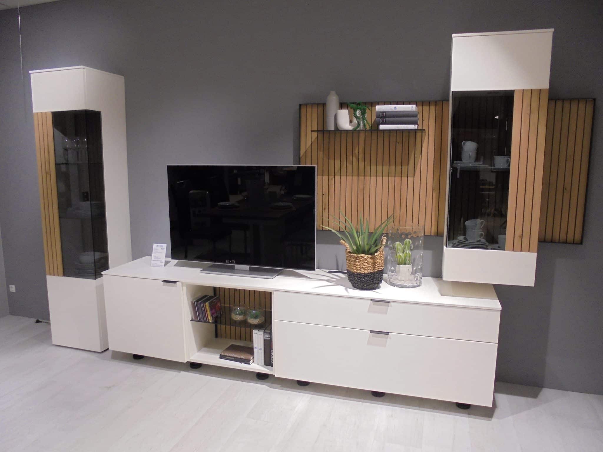 Wohnprogramm Linera TV-Element mit weißen Schränken, Holzakzenten und Pflanzen vor einer grauen Wand.