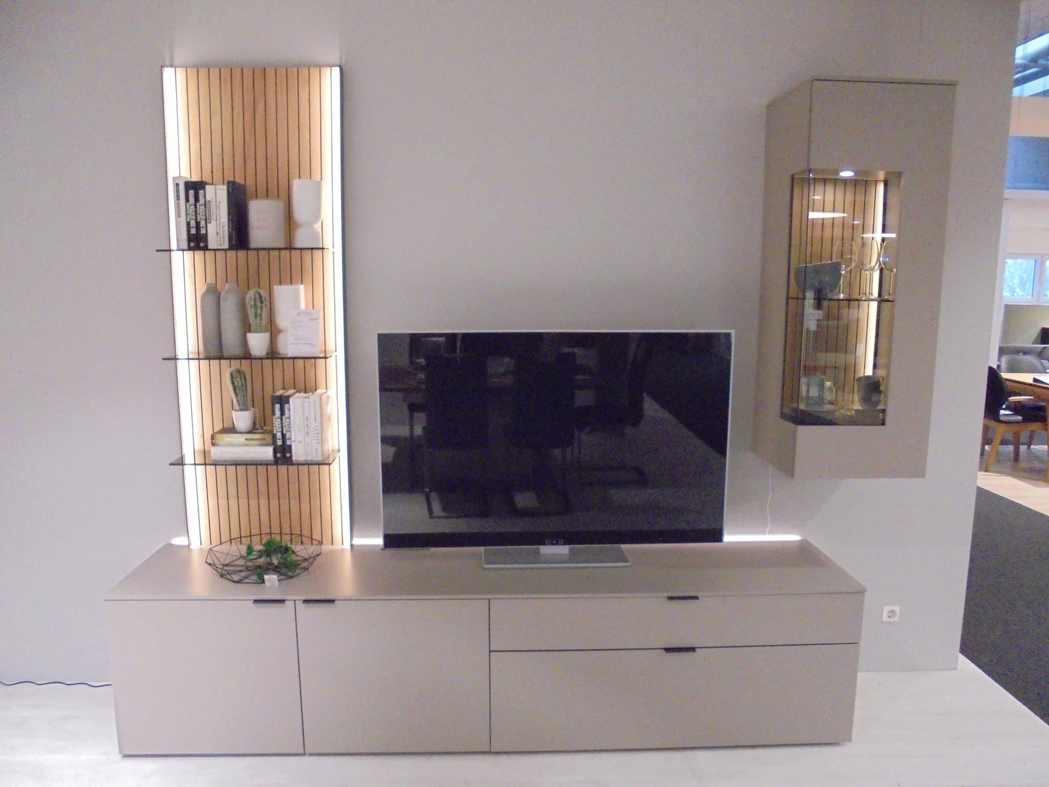 Wohnprogramm Linera: Modernes Wohnzimmer mit TV auf beigem Schrank, offenen Regalen und Glasvitrine an der Wand.