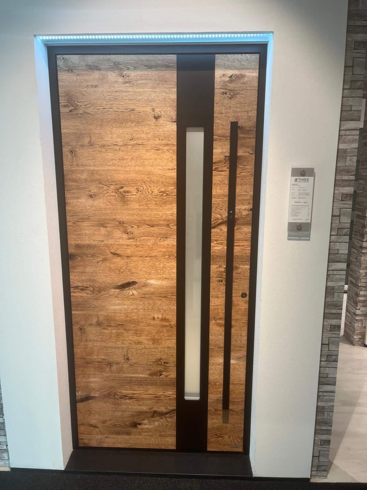 Eine moderne Holztür mit einer vertikalen Glasscheibe und einem Metallgriff, eingefasst in eine weiße Wand mit einem Etikett an der Seite.