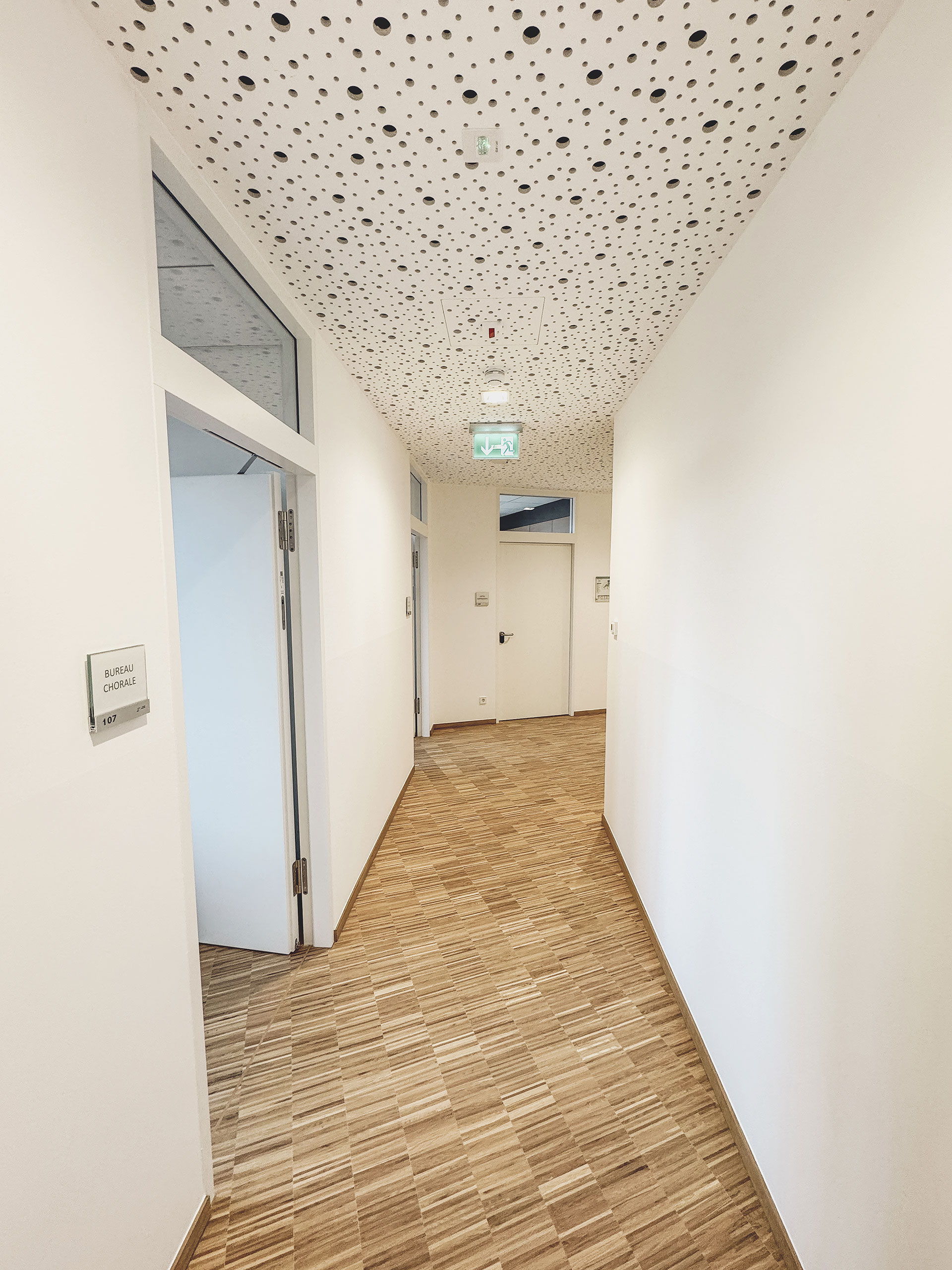 Weißer Flur mit gemusterter Decke, Holzboden und Türen auf beiden Seiten. Am Ende ist ein Ausgangsschild zu sehen.