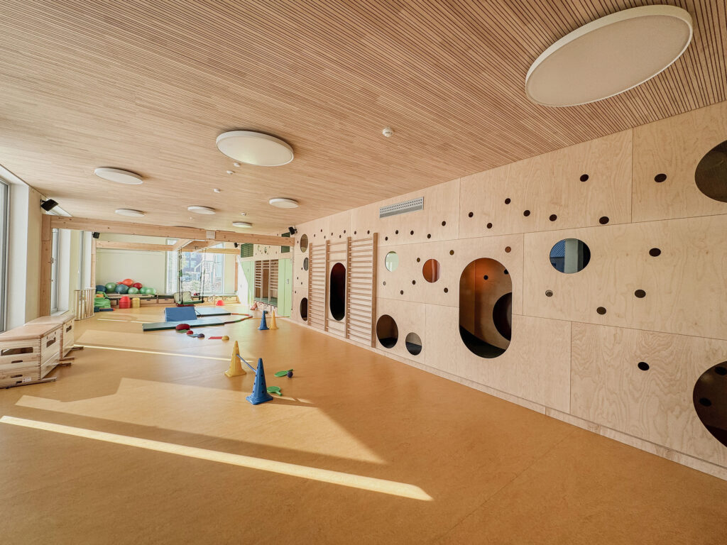 Helles, modernes Indoor-Spielzimmer mit Holzwänden, weichen Spielgeräten und verstreuten Hütchen und Spielsachen auf dem Boden.
