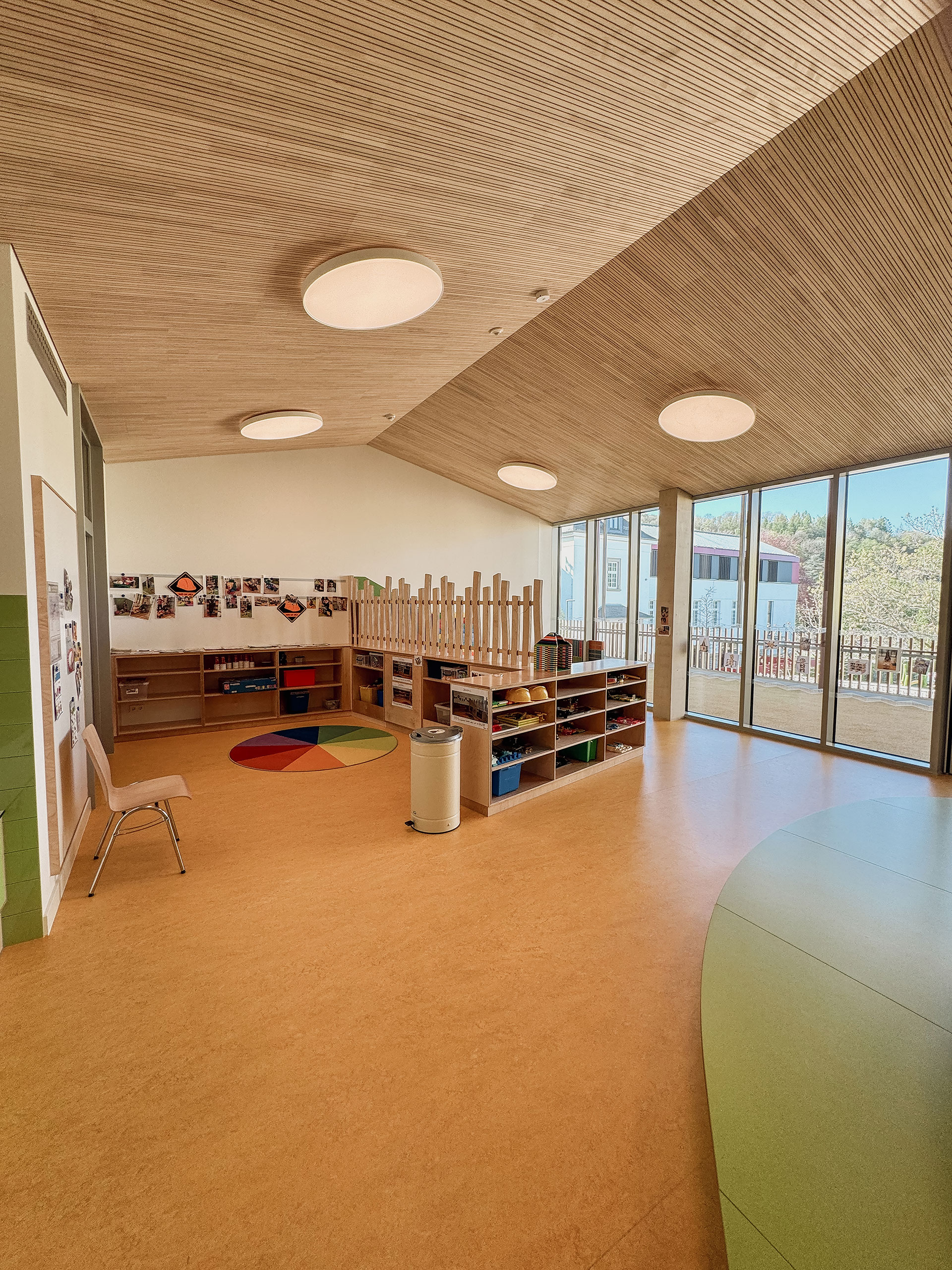 Ein helles, modernes Klassenzimmer mit Holzdecke, großen Fenstern, Regalen, einem Stuhl und bunten Spielmatten auf dem Boden.