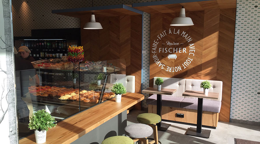 Eine gemütliche Bäckerei mit ausgestelltem Gebäck, kleinen Tischen mit Topfpflanzen und Holzdekor mit Fischer an der Wand.
