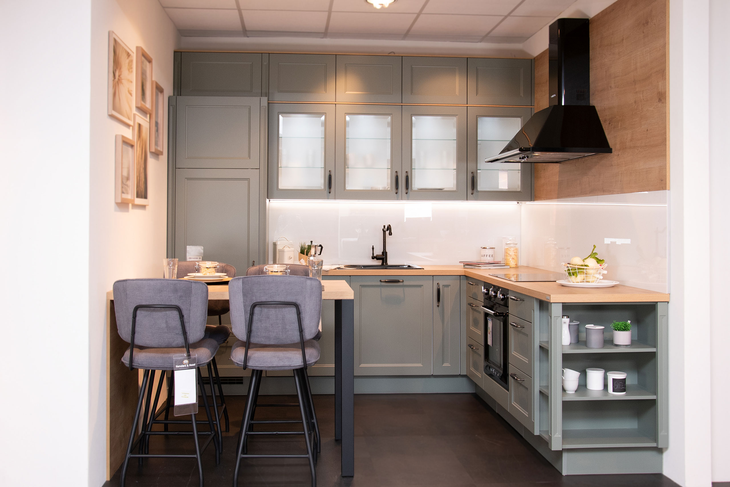 Moderne Küche mit grauen Schränken, Frühstücksbar, zwei Hockern, Einbauofen und offenen Regalen mit Geschirr.