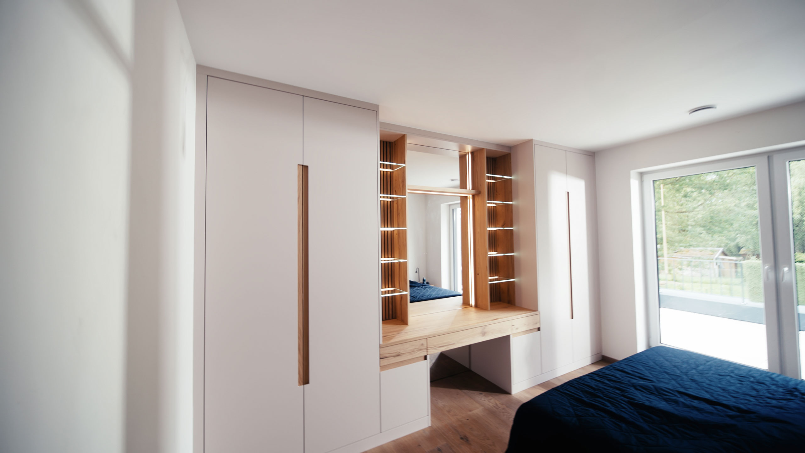 Modernes Schlafzimmer mit weißen Einbauschränken, Holzregalen, einem Kosmetikspiegel und einem großen Fenster, das natürliches Licht hereinlässt.