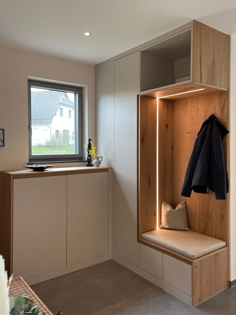 Moderner Eingangsbereich mit eingebauter Holzbank, Kleiderhaken, Kissen, Jacke, Schränken und einem Fenster mit Blick nach draußen.