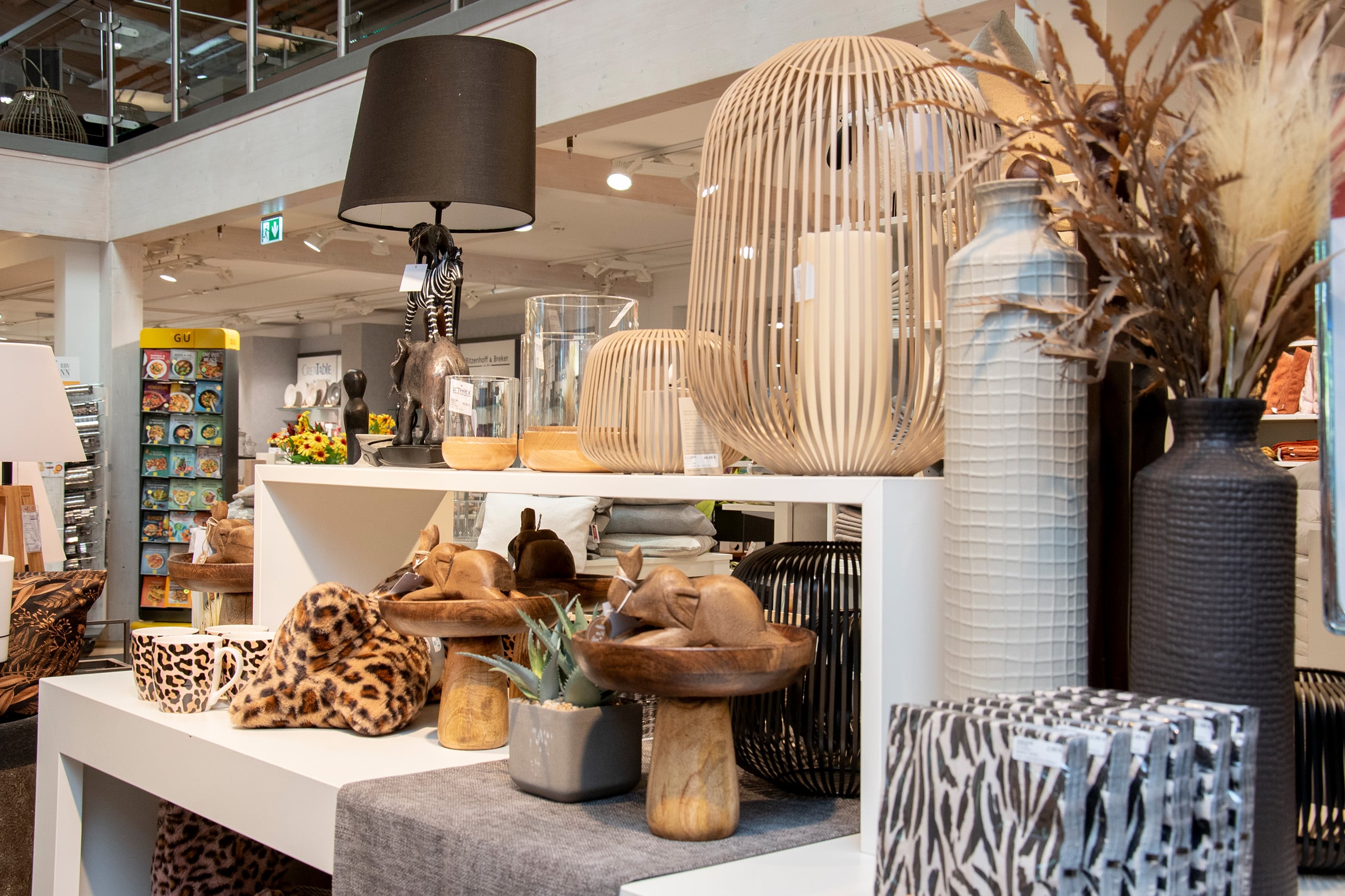 Auslage von dekorativen Wohnaccessoires wie Lampen, Vasen, hölzernen Enten und Artikeln mit Tiermotiven in einem Geschäft.