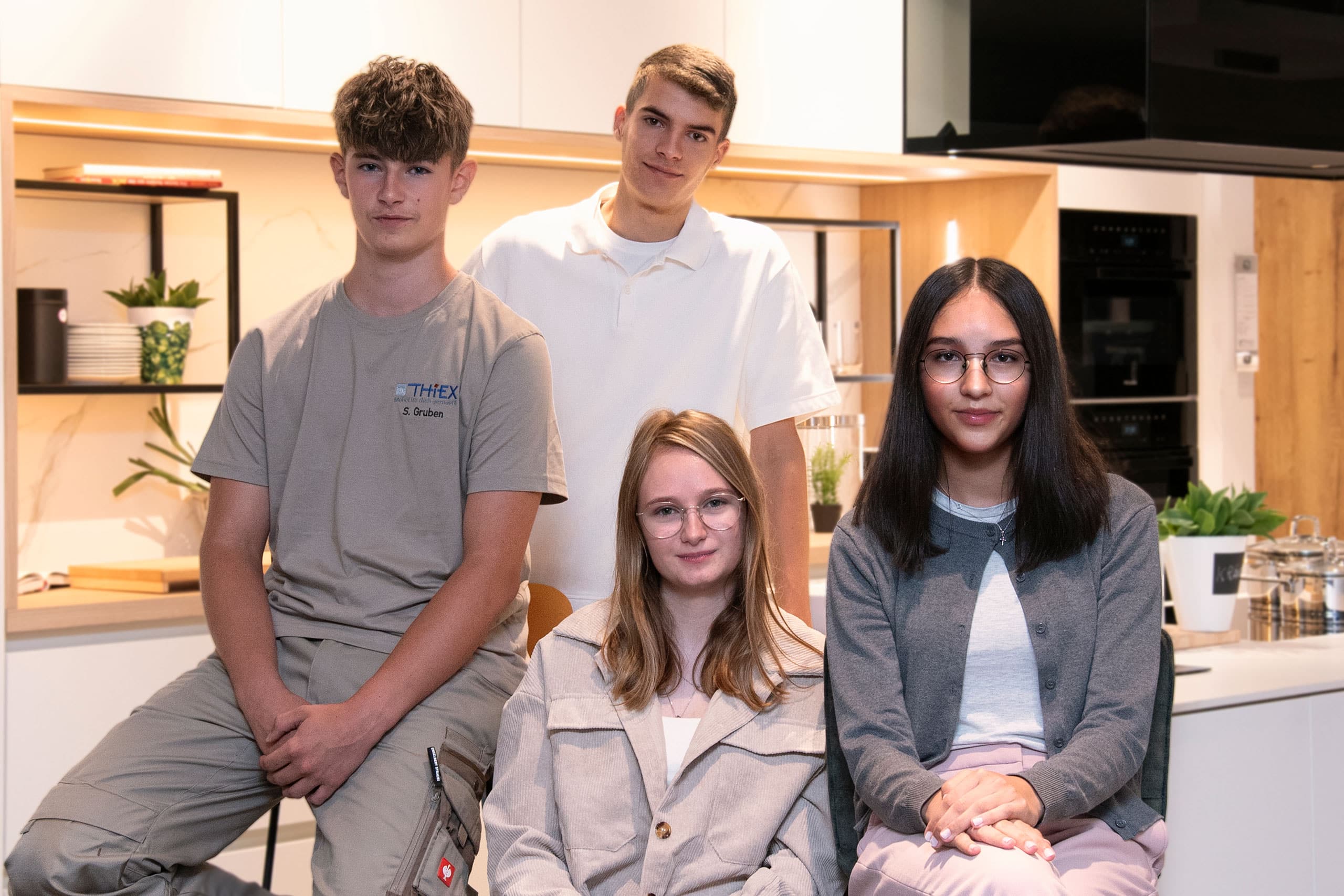 Vier junge Erwachsene posieren gemeinsam in einer modernen Küche, alle tragen Freizeitkleidung und Brillen.