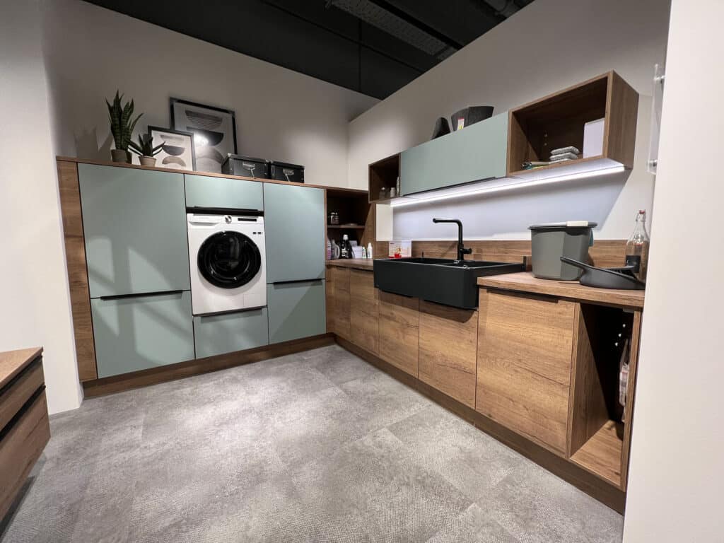 Moderne Waschküche mit Schränken aus Holz und Salbeigrün, einer Waschmaschine, einem schwarzen Waschbecken und verschiedenen Haushaltsgegenständen.