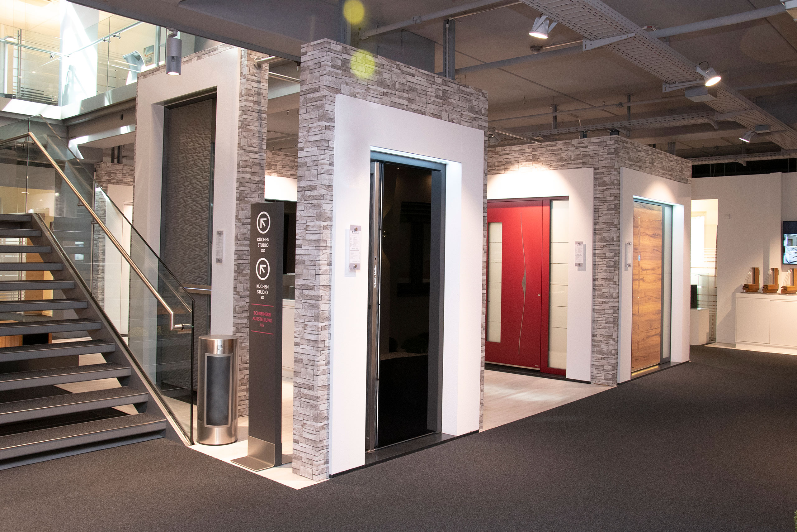 Ausstellungsraum mit modernen Aufzugsdisplays, verschiedenen Türausführungen und einem Treppenhaus auf der linken Seite.