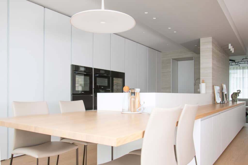 Minimalistische moderne Küche mit hellem Holztisch, weißen Stühlen und schwarzen Einbaugeräten an einer weißen Wand.
