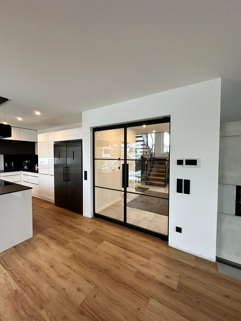 Moderne Küche mit Holzfußboden, schwarzem Kühlschrank, Glasschiebetür und Blick auf die Treppe im Hintergrund.